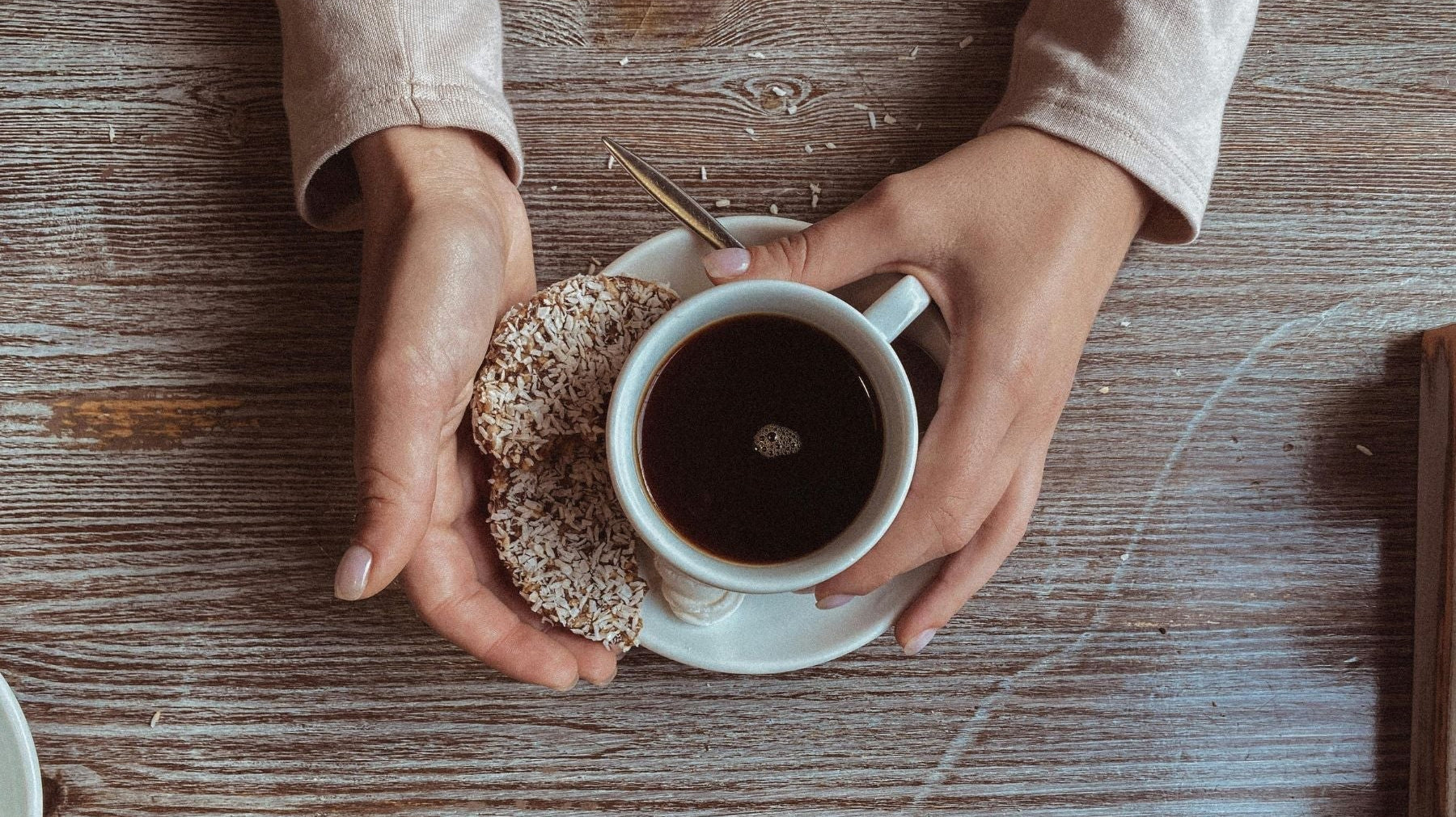 Top view of a cup of black tea with a coconut donut on a wooden table, held in hands during a tea break.