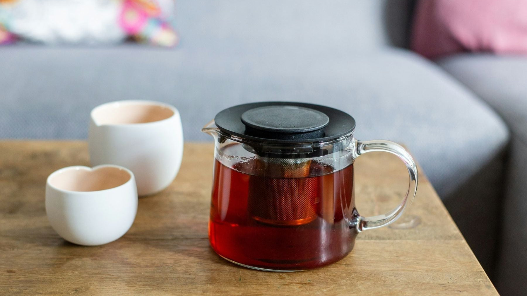 Glass teapot with brewed black tea on a wooden table beside two white cups