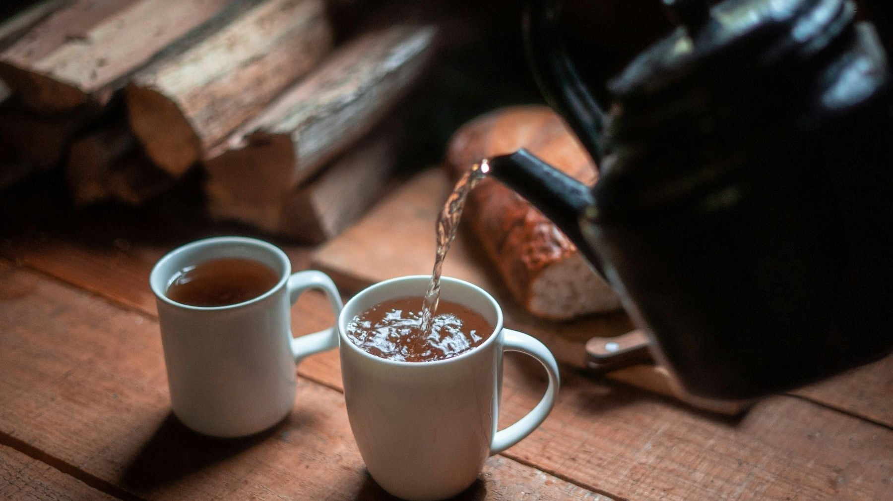 Tea poured into two white mugs on a wooden table beside stacked firewood