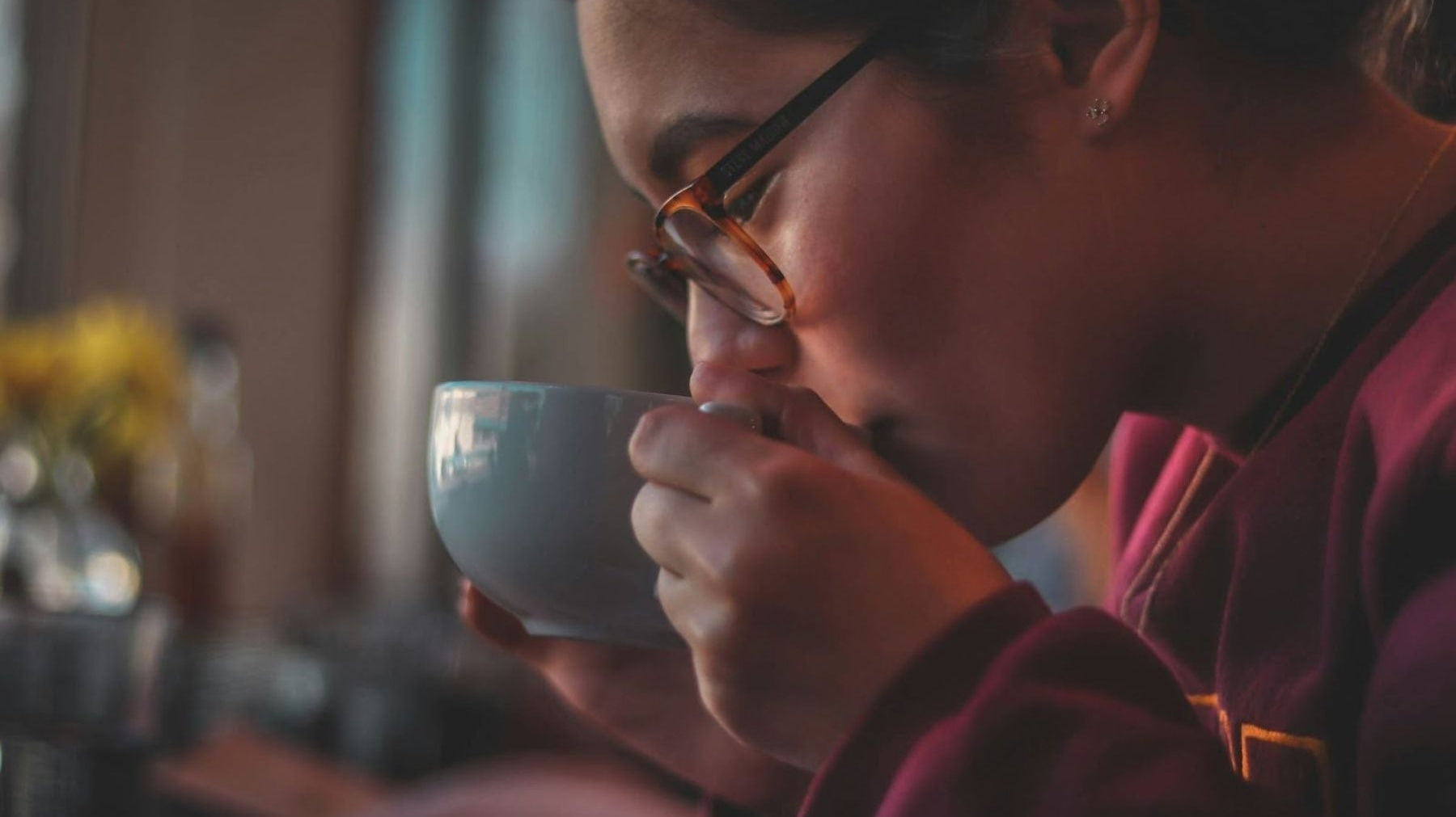 Person wearing glasses sipping tea from a cup in a softly lit setting