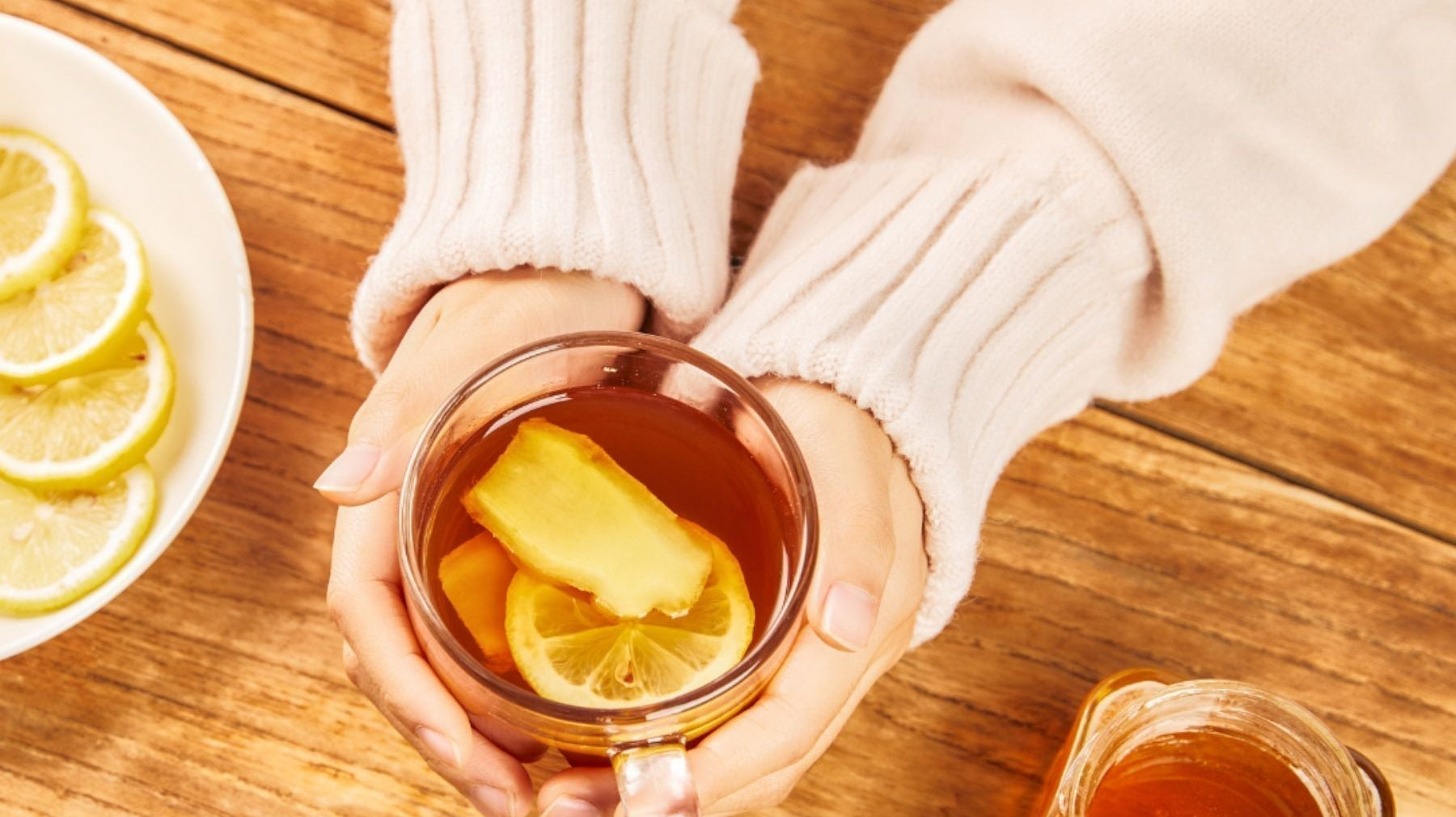 Hands holding a glass mug of tea with lemon and ginger on a wooden table