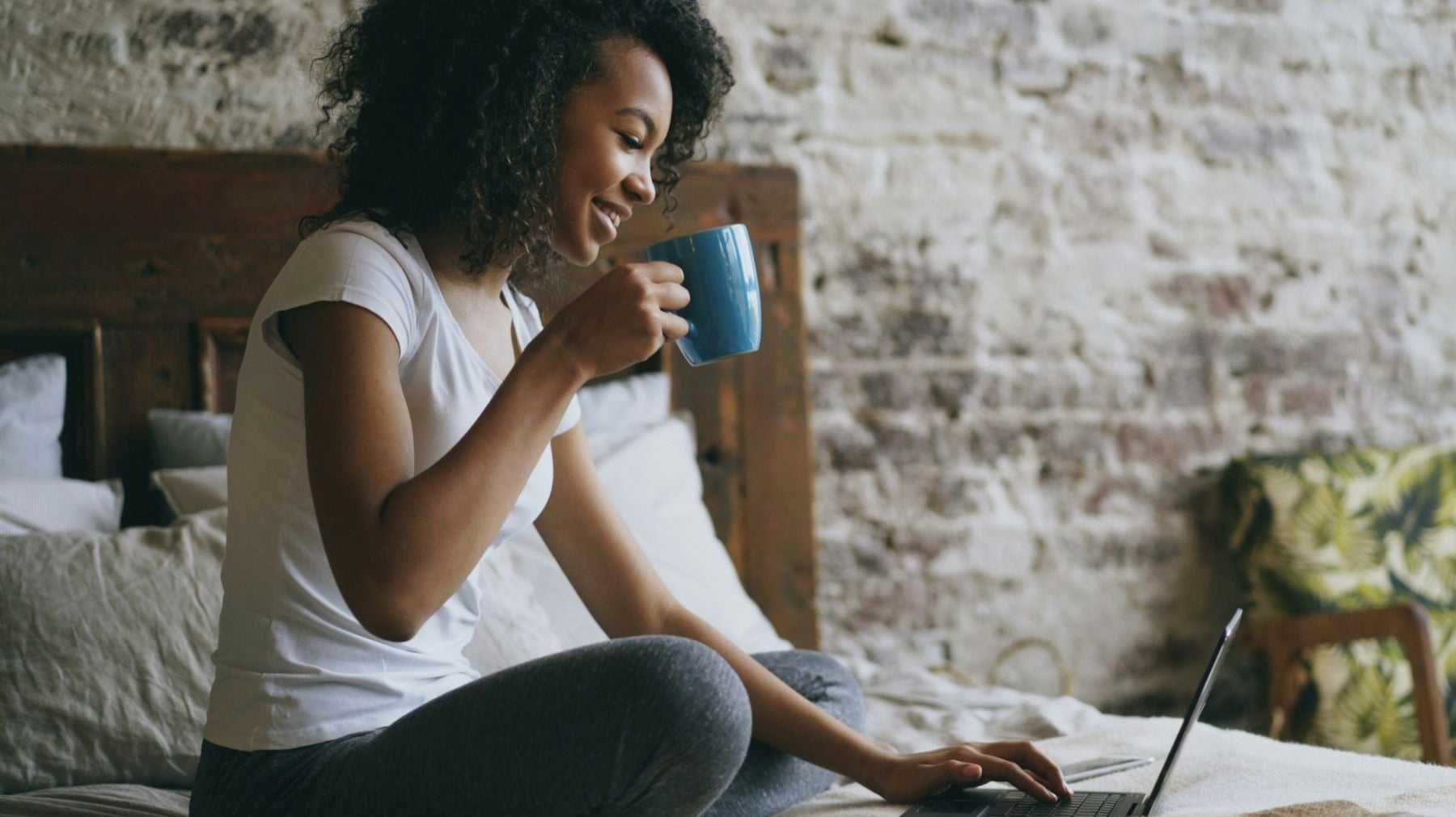 Person sitting on a bed using a laptop while holding a mug of tea