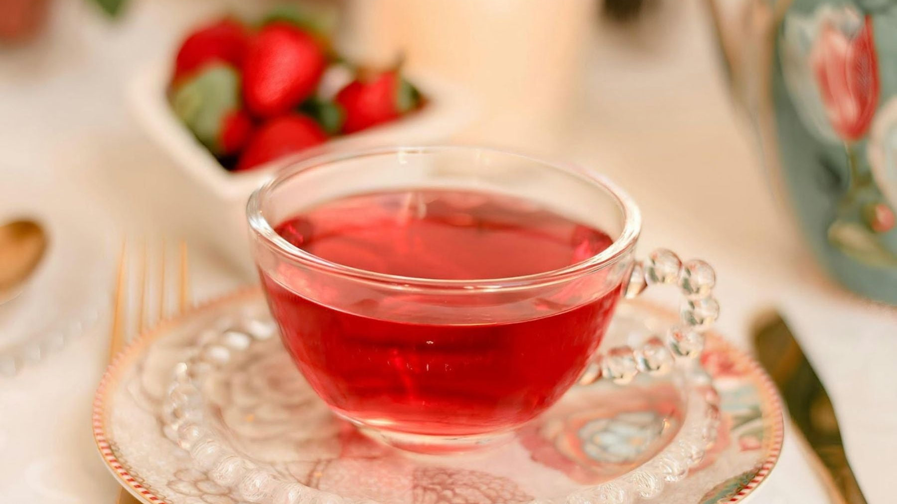 Clear glass cup of red tea on a decorative saucer with strawberries in the background