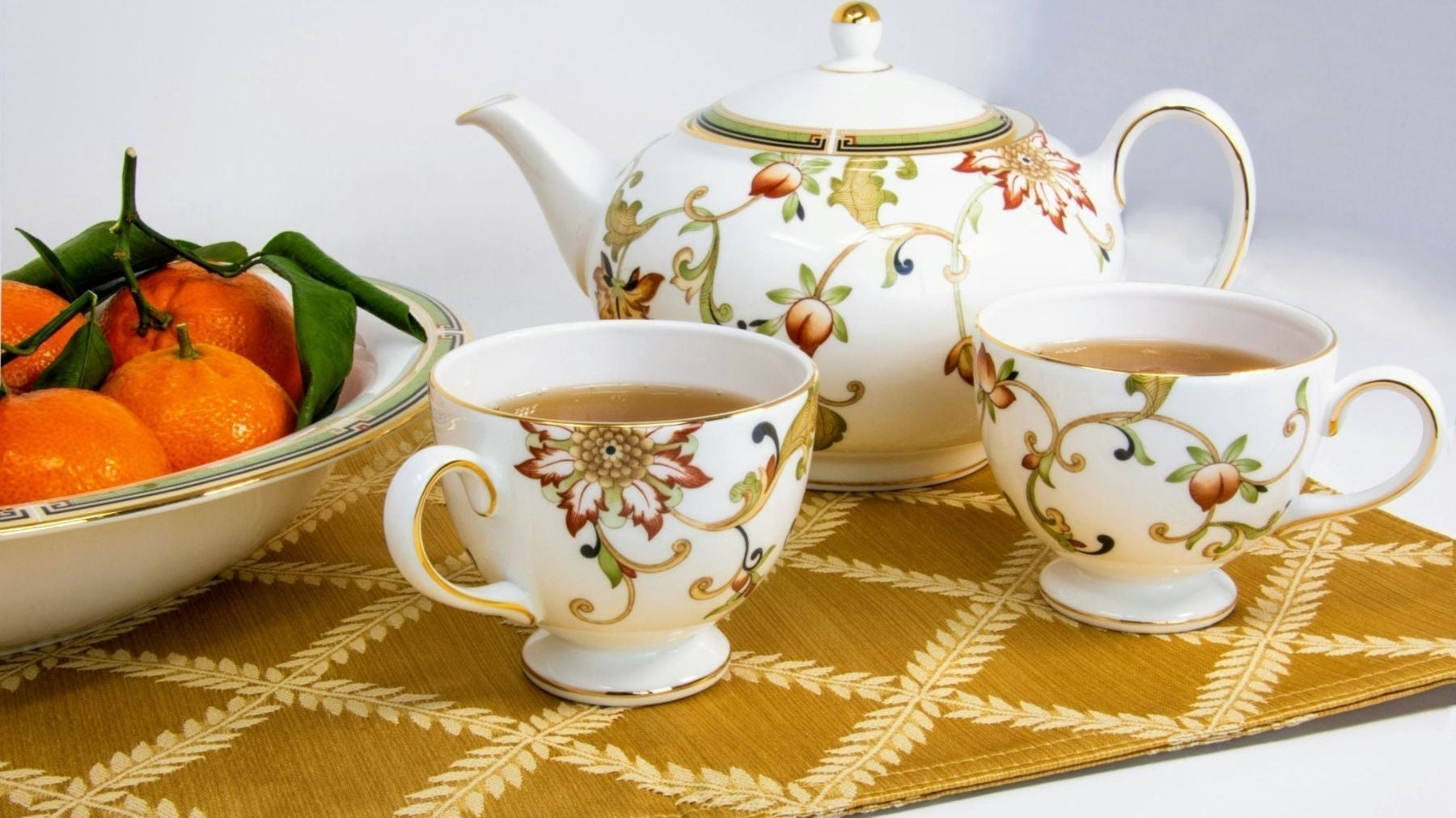 Floral teapot and two teacups filled with tea on a patterned table mat, beside a bowl of fresh oranges