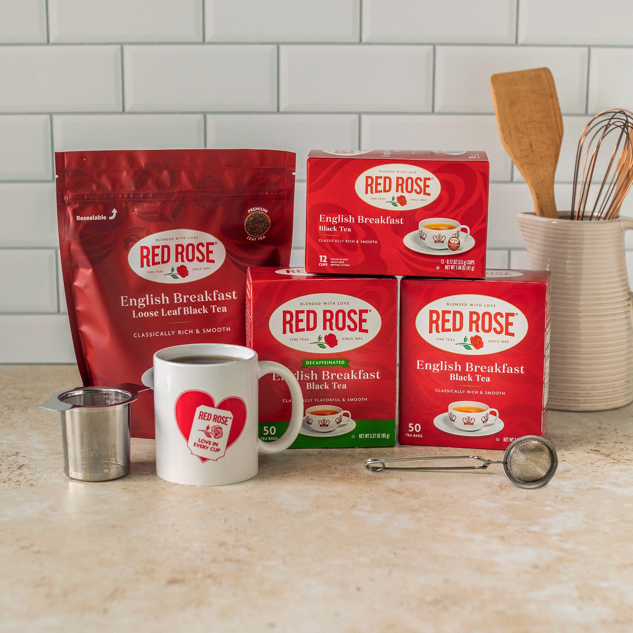 A countertop display of Red Rose English Breakfast black tea products, including a large red bag, three red boxes, a white mug with a red heart logo, a metal tea infuser, and a snapball tea infuser. In the background is a wooden spoon in a ceramic holder against a white subway tile backsplash.