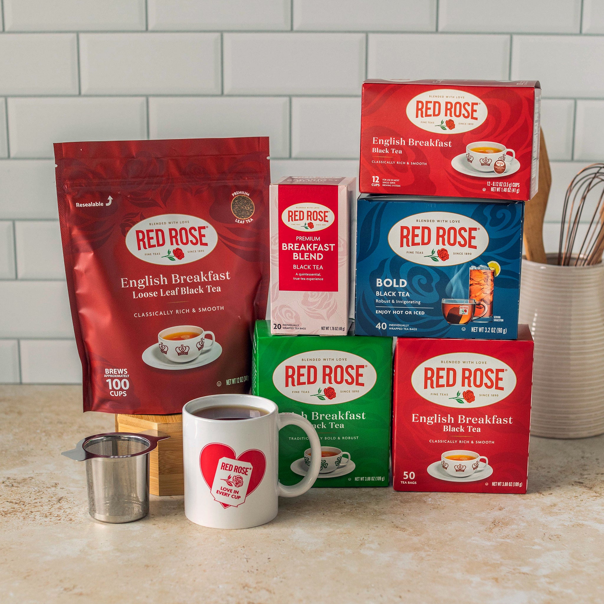 A collection of Red Rose Black Tea products arranged on a kitchen counter in front of a white subway tile backsplash. The products include a large bag of English Breakfast loose leaf black tea, several boxes of tea blends and single-serve tea, and a white ceramic mug with a Red Rose heart logo, along with a small metal strainer.