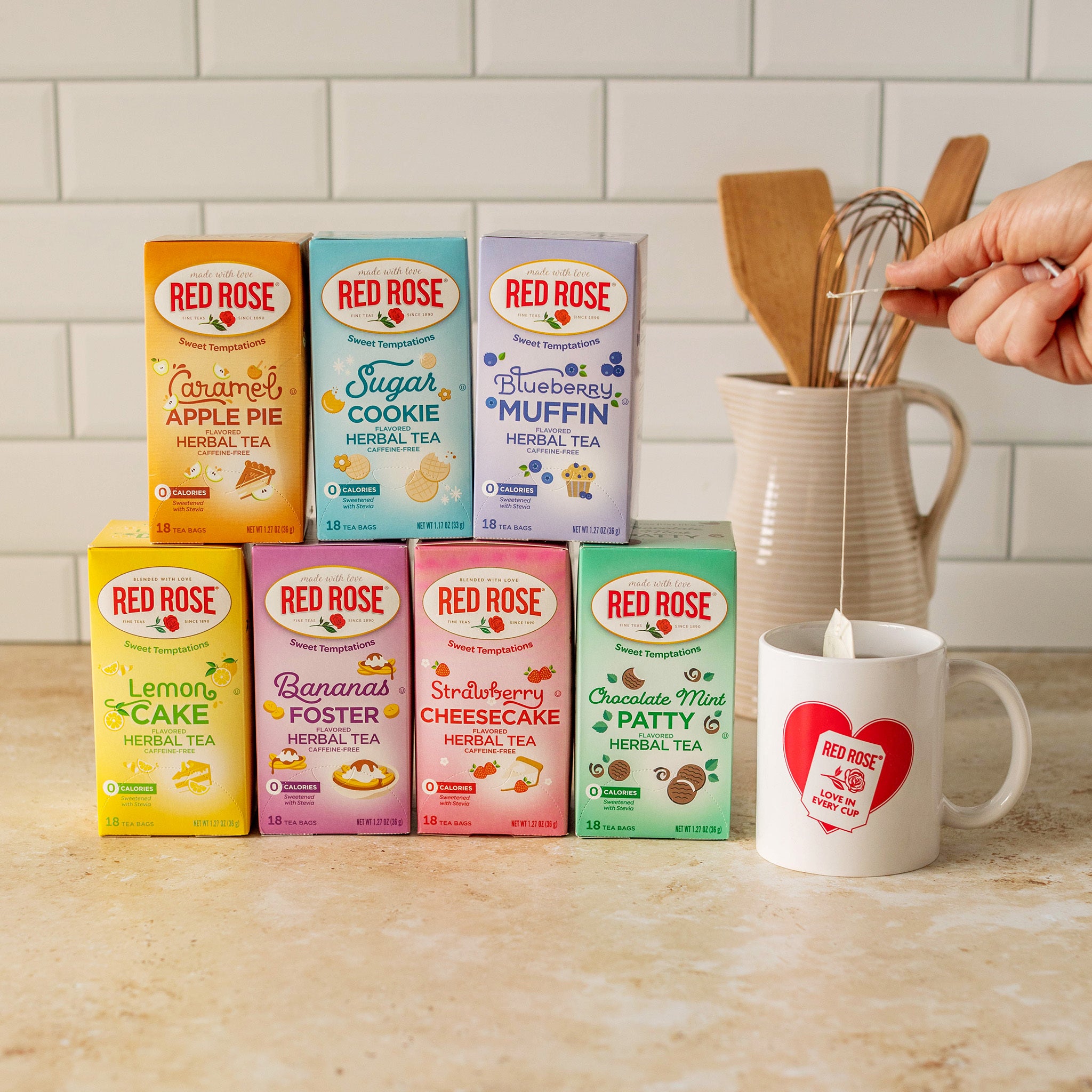 7 Red Rose herbal tea boxes arranged on a kitchen counter, with a white mug featuring a large red heart logo and a hand is holding a tea bag hanging in it. There is a whisk in a ceramic pitcher behind the products in the background