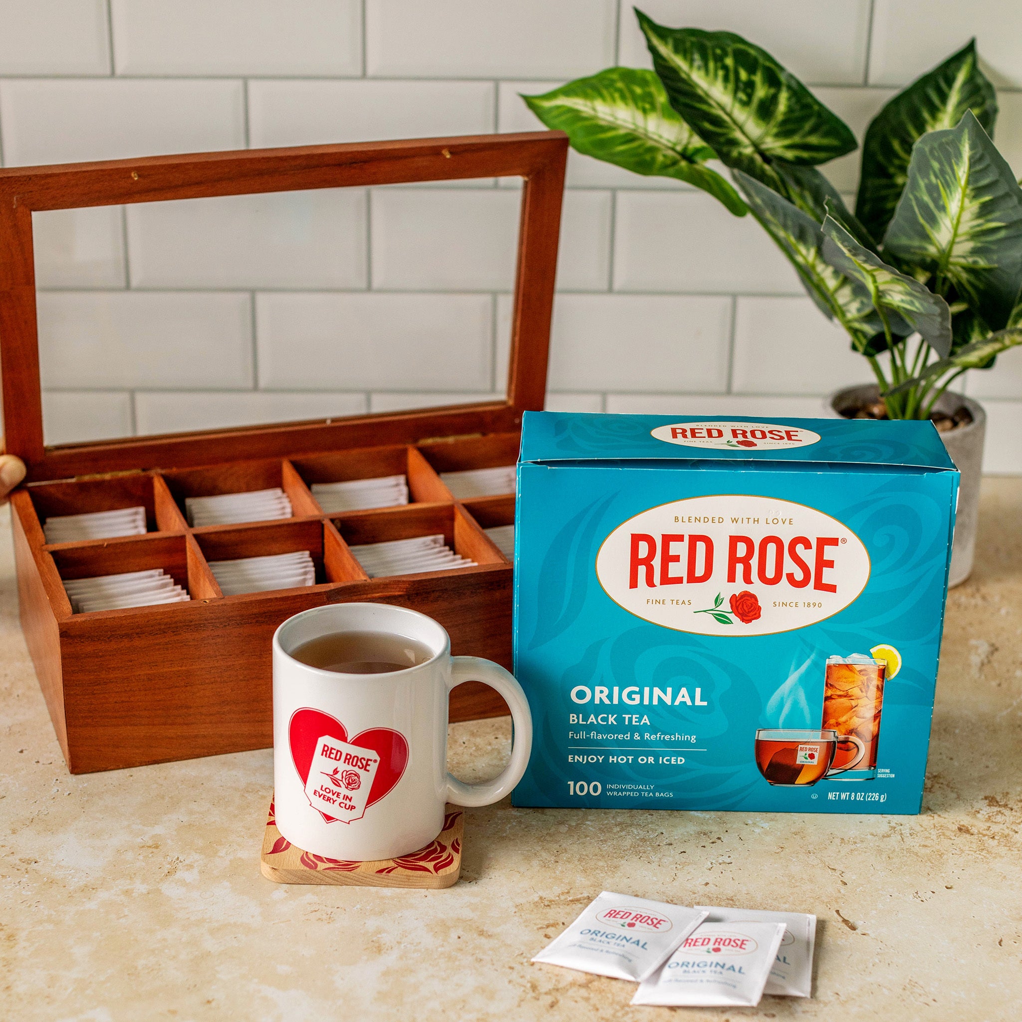 A box of Red Rose Original Black Tea alongside an open wooden tea storage box, a white mug with a Red Rose heart logo, and two tea sachets on a beige countertop with a tiled backsplash and potted plant in the background.