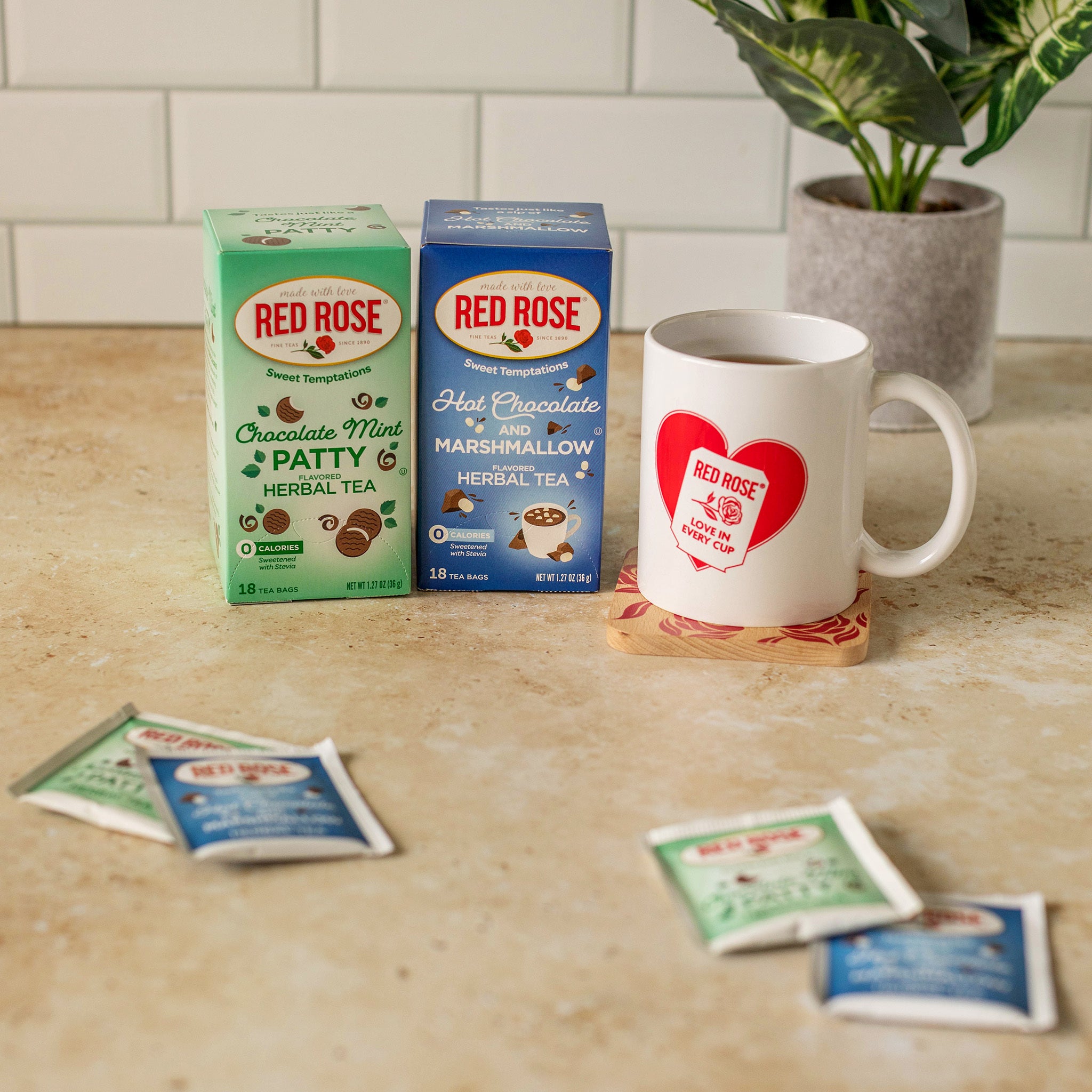 Two Red Rose tea boxes, one Chocolate Mint herbal tea and one Hot Chocolate marshmallow herbal tea, displayed next to a white ceramic mug with a red heart logo. The scene is set on a beige countertop with a tiled backsplash and a potted plant in the background.