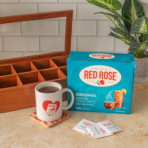Red Rose tea packaging with a mug, coaster, tea chest, and tea bags on a kitchen counter.