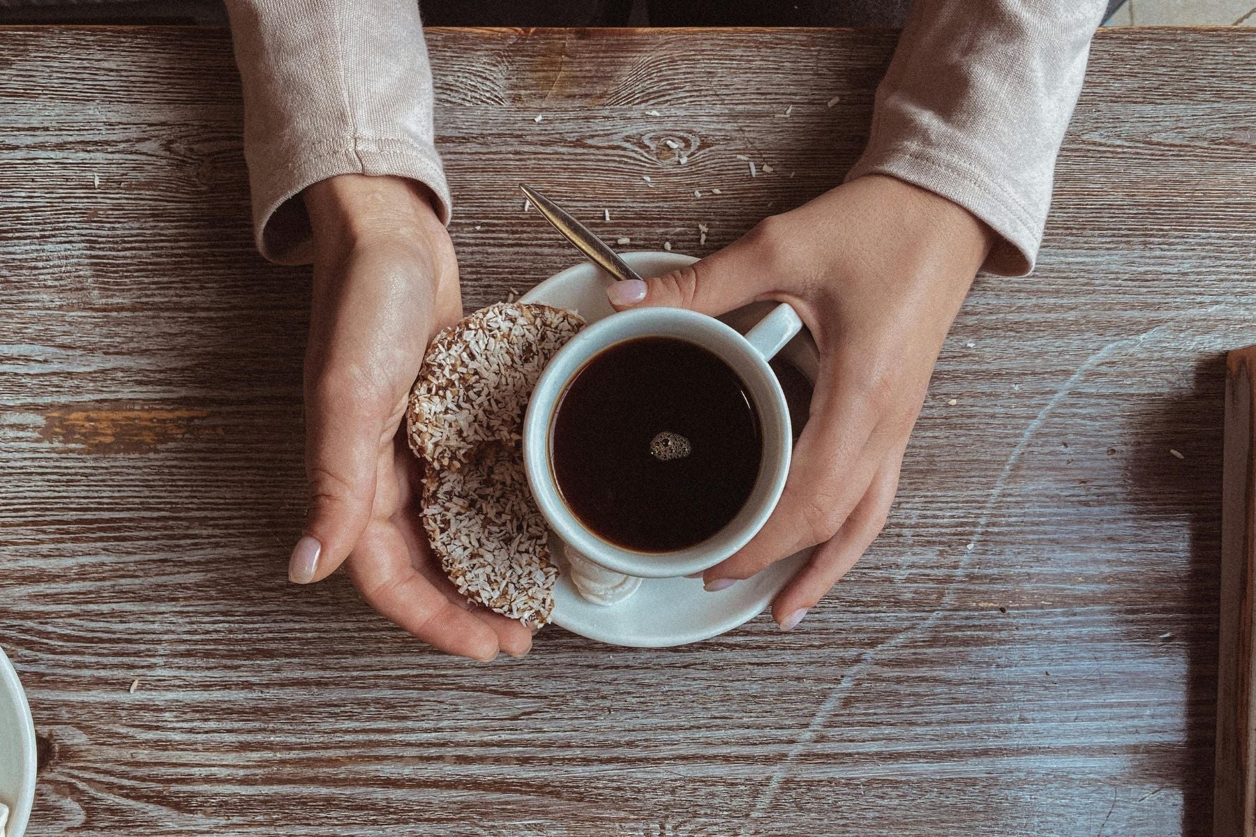 Top view of a cup of black tea with a coconut donut on a wooden table, held in hands during a tea break.
