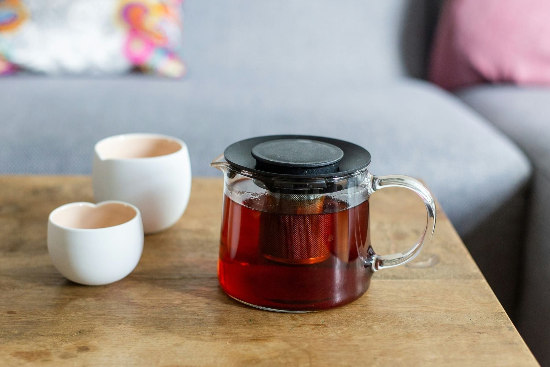 Glass teapot with brewed black tea on a wooden table beside two white cups