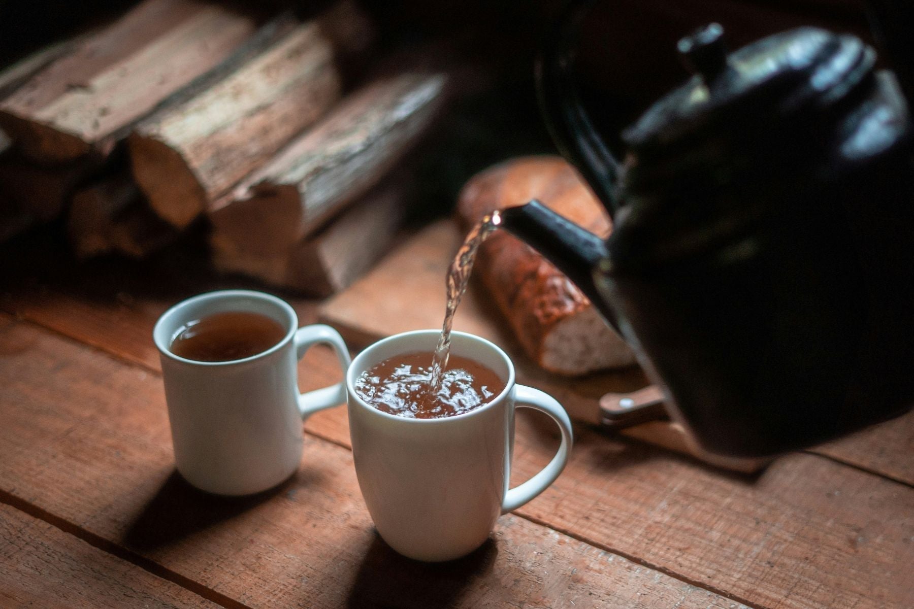 Tea poured into two white mugs on a wooden table beside stacked firewood