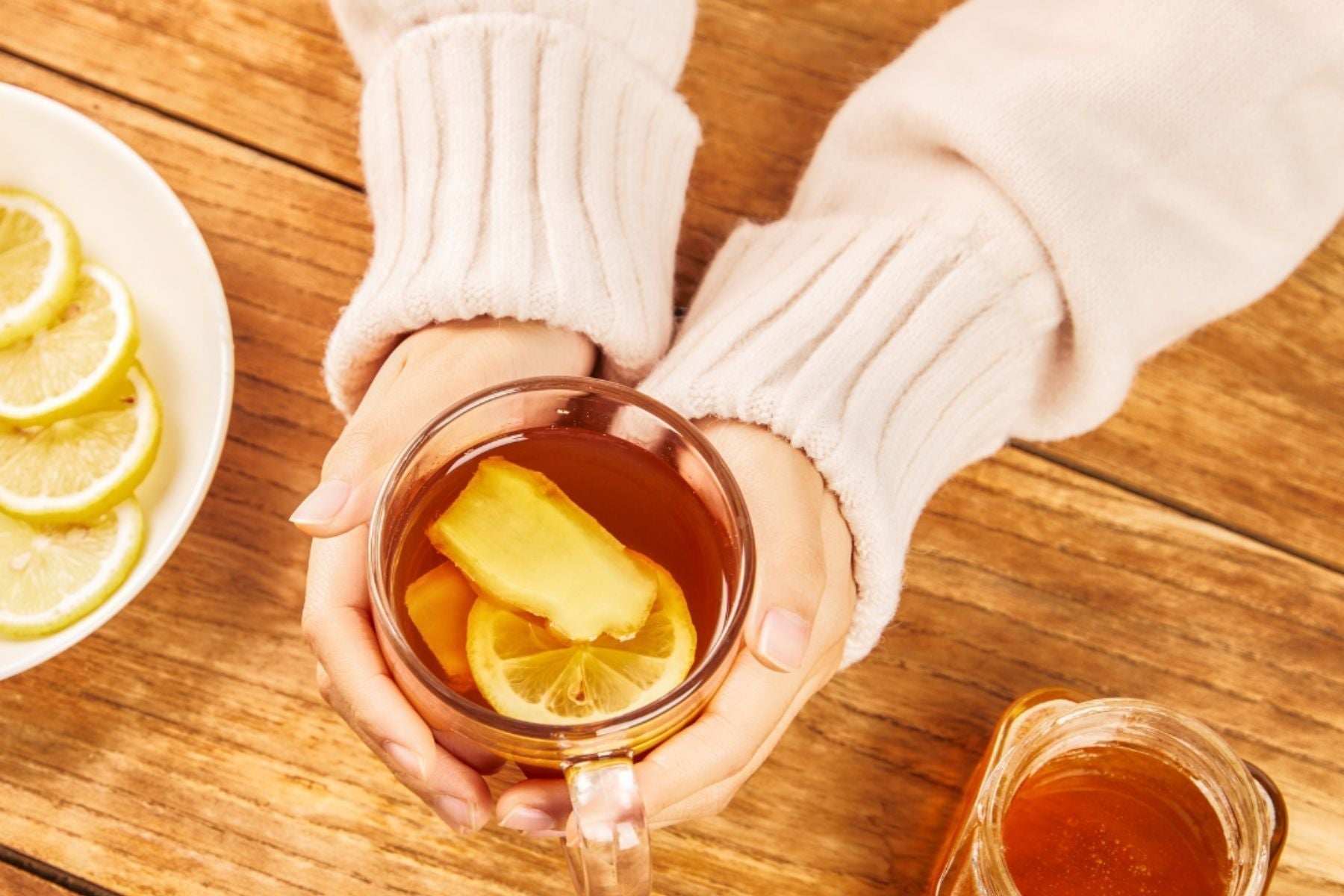 Hands holding a glass mug of tea with lemon and ginger on a wooden table