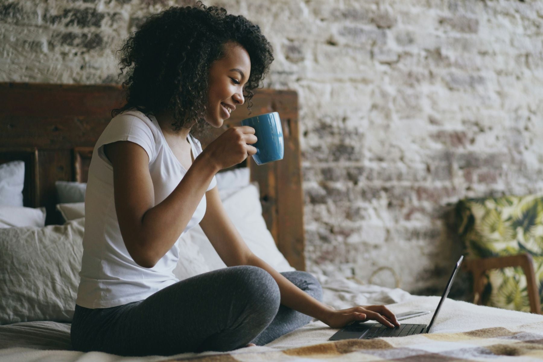Person sitting on a bed using a laptop while holding a mug of tea