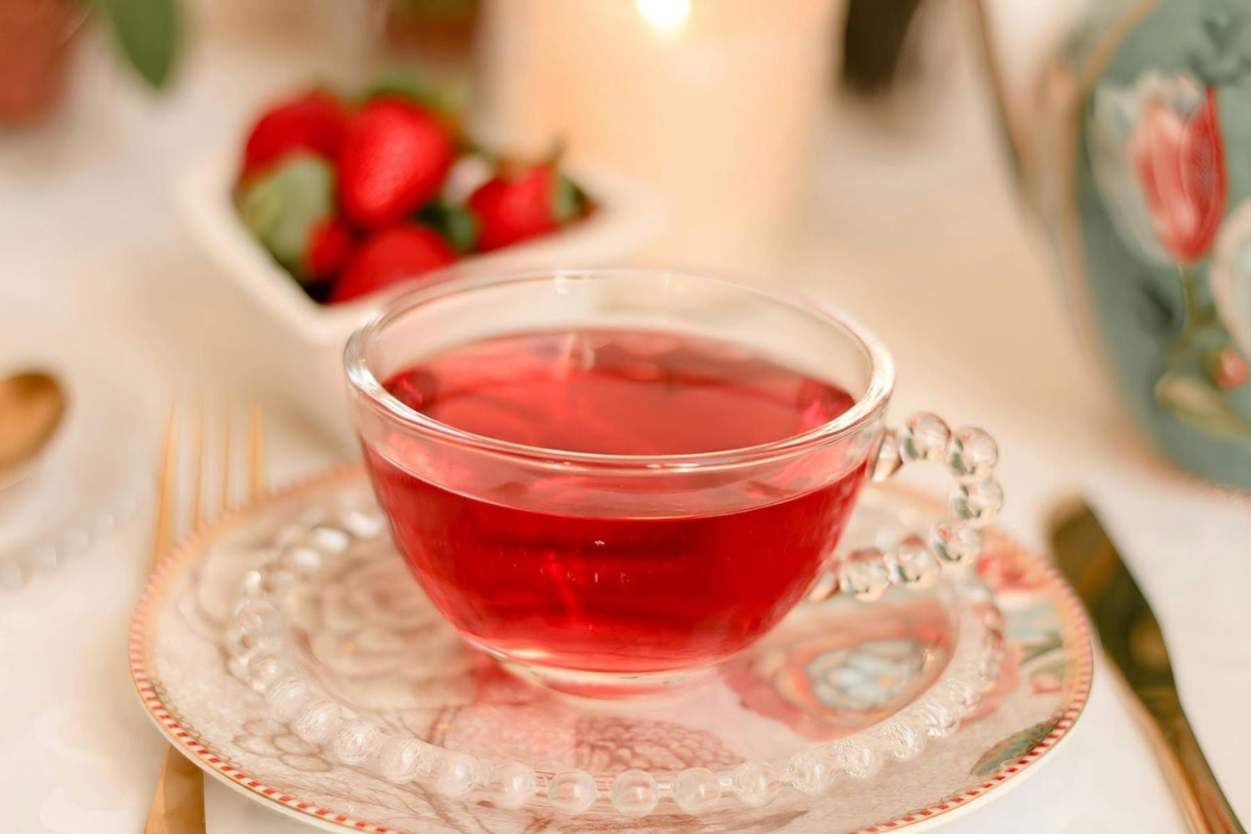 Clear glass cup of red tea on a decorative saucer with strawberries in the background