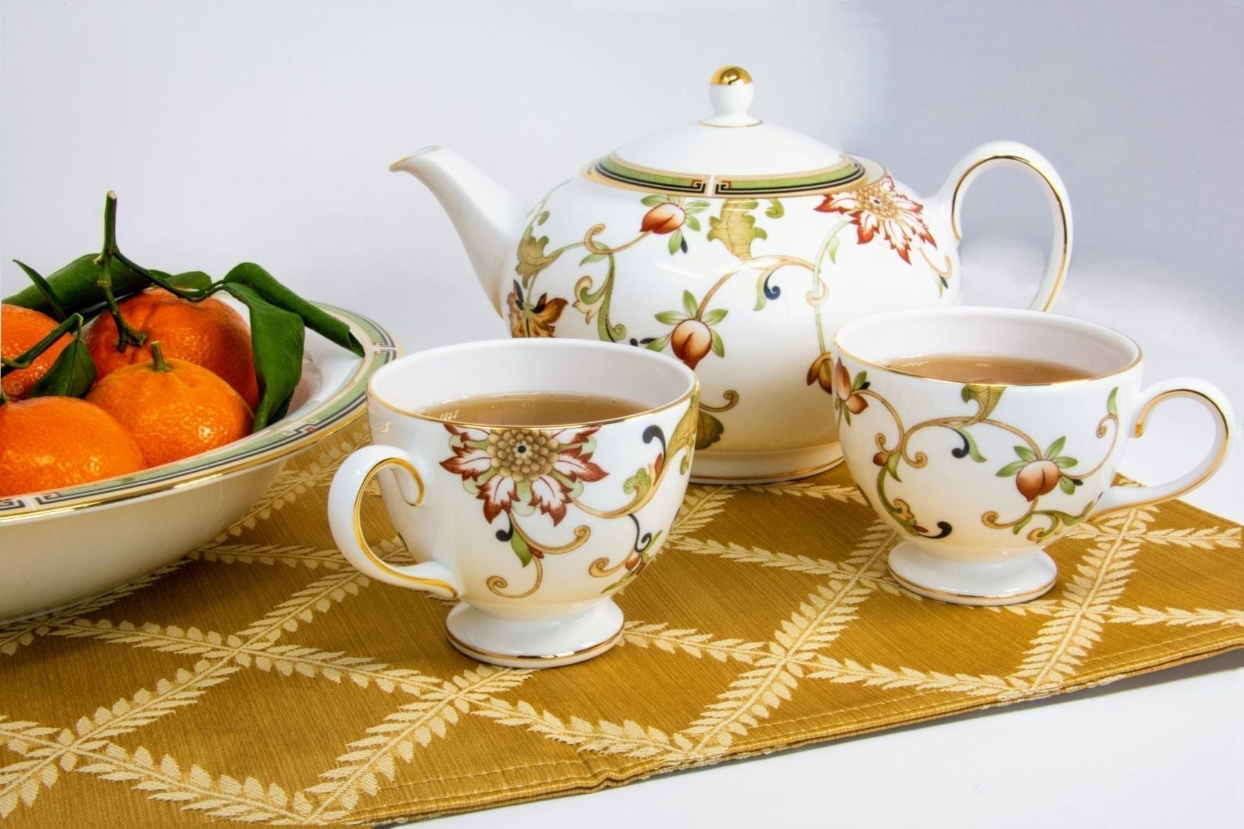 Floral teapot and two teacups filled with tea on a patterned table mat, beside a bowl of fresh oranges