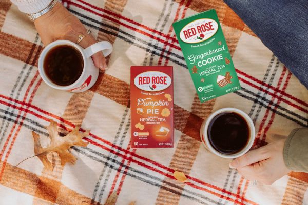 Three Red Rose tea boxes arranged on a plaid blanket with two white mugs of black coffee and a hand visible holding one mug. The visible boxes are Pumpkin Pie Herbal Tea, Gingerbread Cookie Herbal Tea, and Red Rose Gingerbread Cookie Mocha Tea.