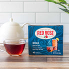 Box of Red Rose Bold Black Tea with a cup of tea on a wooden surface.
