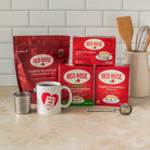 A countertop display of Red Rose English Breakfast black tea products, including a large red bag, three red boxes, a white mug with a red heart logo, a metal tea infuser, and a snapball tea infuser. In the background is a wooden spoon in a ceramic holder against a white subway tile backsplash.