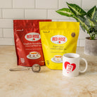 Two Red Rose loose leaf tea bags, one in a red pouch and one in a yellow pouch, displayed on a beige countertop next to a white ceramic mug with a red heart logo and a metal tea strainer. A potted green plant is visible in the background against a white subway tile wall.