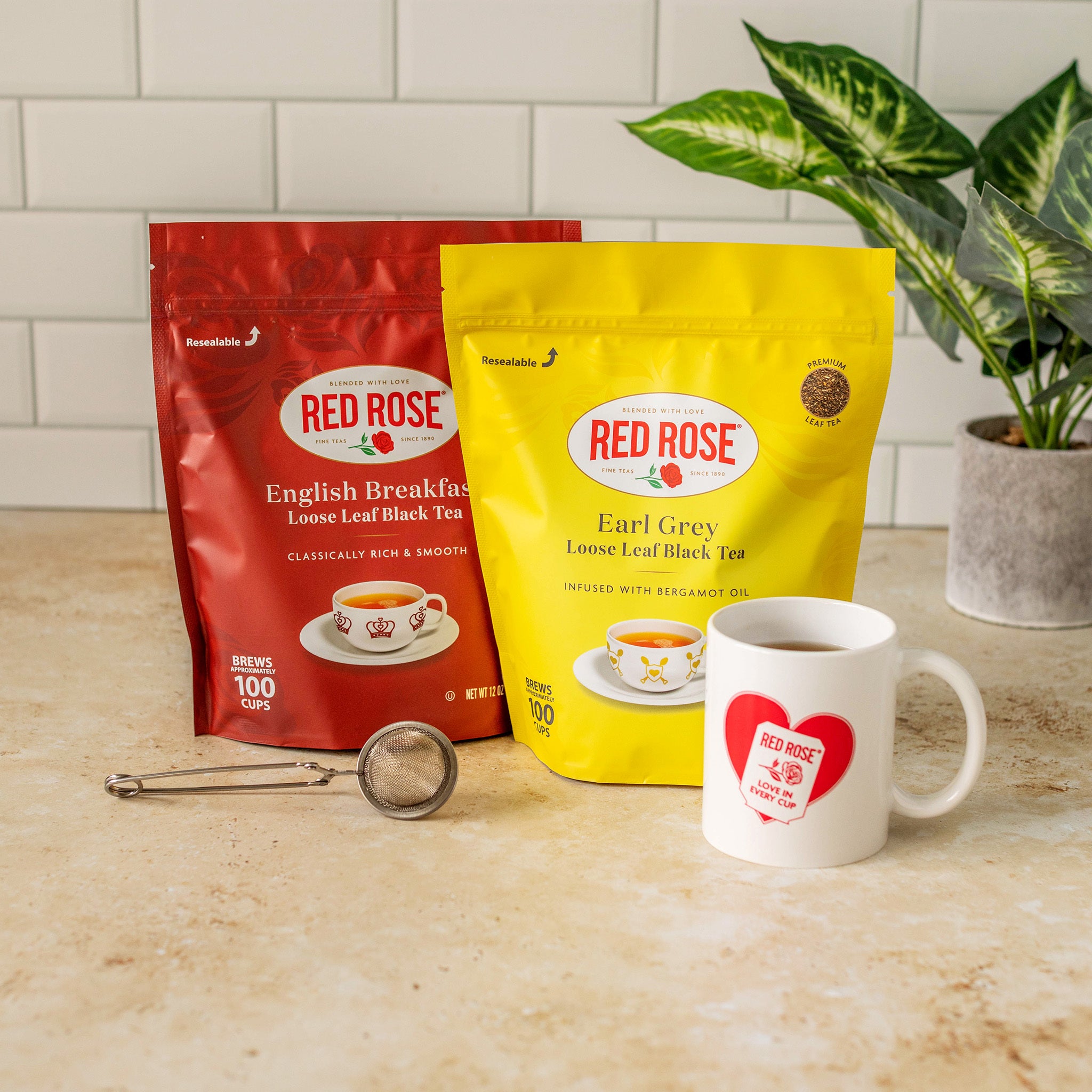 Two Red Rose loose leaf tea bags, one in a red pouch and one in a yellow pouch, displayed on a beige countertop next to a white ceramic mug with a red heart logo and a metal tea strainer. A potted green plant is visible in the background against a white subway tile wall.