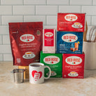 A collection of Red Rose Black Tea products arranged on a kitchen counter in front of a white subway tile backsplash. The products include a large bag of English Breakfast loose leaf black tea, several boxes of tea blends and single-serve tea, and a white ceramic mug with a Red Rose heart logo, along with a small metal strainer.