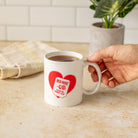 A hand holds a white ceramic mug with a red heart graphic that reads 'RED ROSE LOVE IN EVERY CUP' on a beige countertop in a kitchen setting.