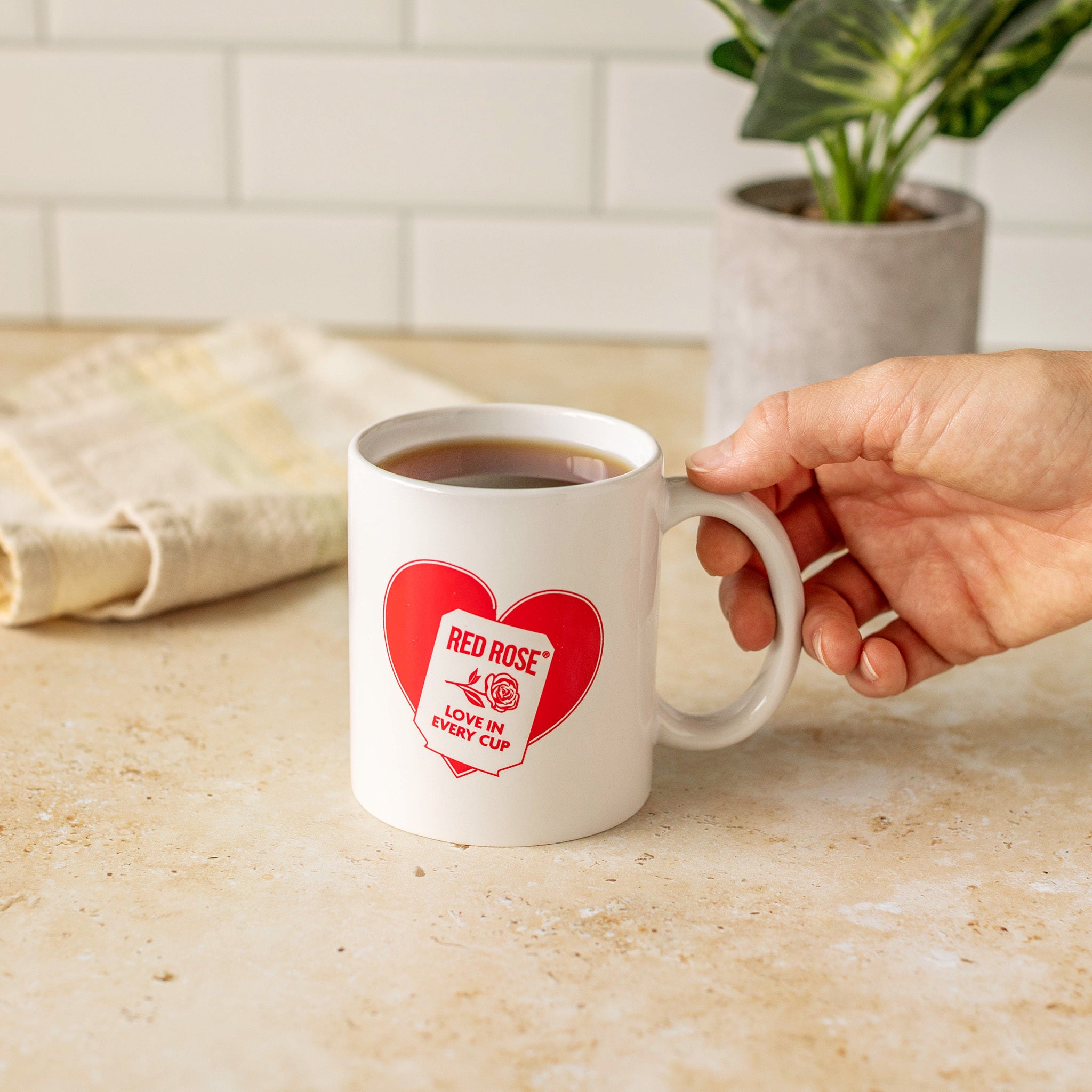 A hand holds a white ceramic mug with a red heart graphic that reads 'RED ROSE LOVE IN EVERY CUP' on a beige countertop in a kitchen setting.