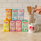 7 Red Rose herbal tea boxes arranged on a kitchen counter, with a white mug featuring a large red heart logo and a hand is holding a tea bag hanging in it. There is a whisk in a ceramic pitcher behind the products in the background