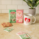 Two Red Rose herbal tea boxes standing on a countertop, next to a white ceramic mug with a red heart and Red Rose label, with a few tea bags scattered in front and a potted plant in the background.
