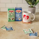 Two Red Rose tea boxes, one Chocolate Mint herbal tea and one Hot Chocolate marshmallow herbal tea, displayed next to a white ceramic mug with a red heart logo. The scene is set on a beige countertop with a tiled backsplash and a potted plant in the background.