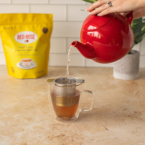 Red teapot pouring tea into a glass teacup with a tea infuser on a kitchen counter with a yellow Red Rose tea bag in the background.