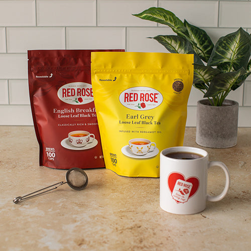 Red Rose loose leaf tea blends, a tea infuser, and a mug on a countertop with a plant in the background.