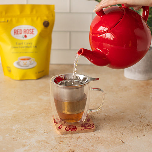 Red teapot pouring tea into a glass mug with a tea infuser on a countertop with a Red Rose tea package in the background.