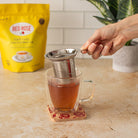 Person using a tea strainer over a cup of tea with Red Rose tea packaging in the background.
